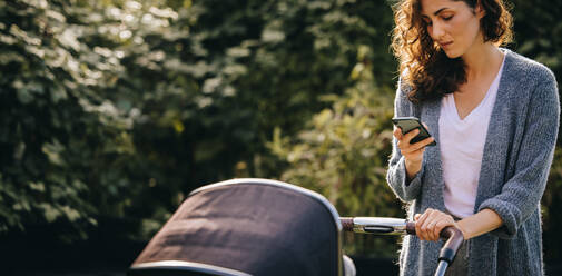 Eine Mutter, die ihr Baby im Kinderwagen durch die Gegend schiebt. Eine Frau, die Nachrichten auf ihrem Smartphone abruft, während sie einen Kinderwagen schiebt. - JLPSF08204