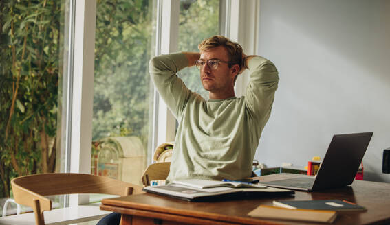 Man sitting at table with hands behind head. Man taking break while working from home. - JLPSF08215