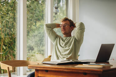 Man sitting at table with eyes closed and hands behind head at home. Man taking break from work. - JLPSF08216