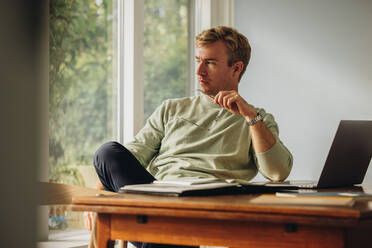 Man sitting at home holding eyeglasses looking away and thinking. Man working at home with laptop in table. - JLPSF08217