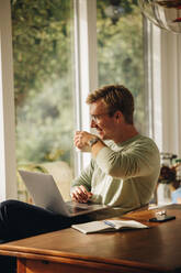 Mann trinkt Kaffee bei der Arbeit von zu Hause aus. Mann sitzt am Tisch mit Laptop und trinkt Kaffee, schaut weg und lächelt. - JLPSF08223