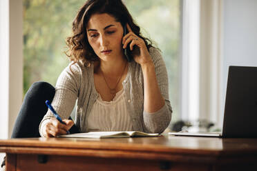 Geschäftsfrau macht sich Notizen, während sie mit dem Handy telefoniert. Frau arbeitet von zu Hause aus. - JLPSF08228