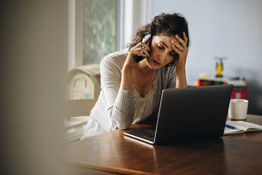 Business woman holding her head and talking on cell phone. Stressed female making a phone call while working on laptop at home. - JLPSF08230