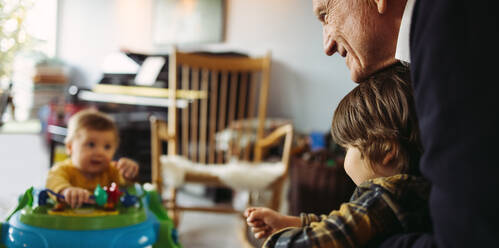 Älterer Mann hält seinen Enkel und betrachtet das Baby, das zu Hause auf dem Rollator sitzt. Großvater mit Enkelkindern zu Hause. - JLPSF08248