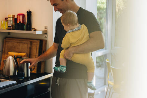 Mann mit Baby, der sich in der Küche einen Kaffee kocht. Mann im Vaterschaftsurlaub mit Baby in der Küche. - JLPSF08256