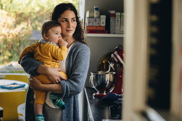 Mutter und Kind zusammen in der Küche mit Blick aus dem Fenster. Mutter trägt ihr Baby zu Hause stehend. - JLPSF08260
