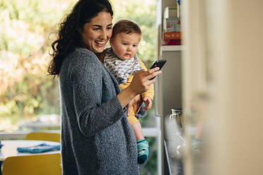 Eine Frau schreibt eine SMS auf ihrem Mobiltelefon, während sie mit ihrem Baby steht. Eine Mutter benutzt ein Mobiltelefon, während sie ihr Kind zu Hause trägt. - JLPSF08262