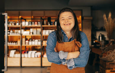 Happy woman with Down syndrome smiling while standing at the entrance of a deli. Empowered woman with an intellectual disability working as a shopkeeper in a local supermarket. - JLPSF08269