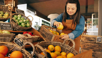 Frau mit Down-Syndrom beim Auffüllen von frischem Obst in der Gemüseabteilung eines Lebensmittelgeschäfts. Selbstbewusste Frau mit geistiger Behinderung bei der Arbeit in einem örtlichen Feinkostladen. - JLPSF08271