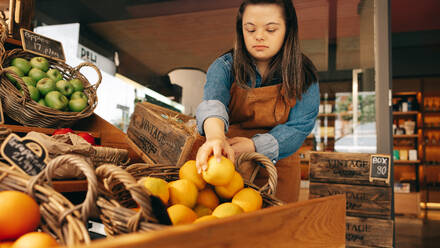 Mitarbeiterin eines Lebensmittelgeschäfts mit Down-Syndrom beim Auffüllen von frischem Obst in der Gemüseabteilung eines Lebensmittelgeschäfts. Selbstständige Frau mit geistiger Behinderung in einem örtlichen Supermarkt. - JLPSF08273