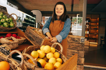 Glückliche Frau mit Down-Syndrom, die lächelnd in der Gemüseabteilung eines Lebensmittelgeschäfts steht. Selbstbewusste Frau mit einer geistigen Behinderung, die in einem örtlichen Supermarkt arbeitet. - JLPSF08274