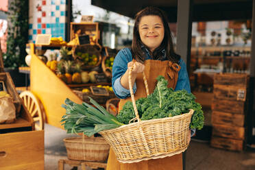 Supermarket employee with Down syndrome holding a basket of fresh organic vegetables in a grocery store. Happy woman with an intellectual disability working in a local convenience store. - JLPSF08276