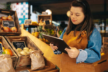 Verkäuferin mit Down-Syndrom bei der Arbeit mit einem digitalen Tablet in der Frischwarenabteilung eines Lebensmittelgeschäfts. Frau mit geistiger Behinderung bei der Arbeit in einem örtlichen Supermarkt. - JLPSF08280