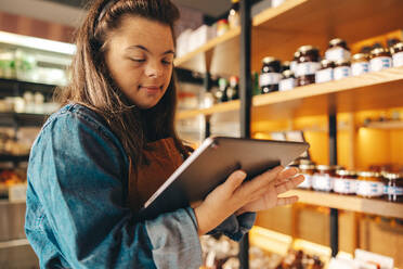 Ladenangestellte mit Down-Syndrom bei der Inventur mit Hilfe eines digitalen Tablets. Selbstständige Frau mit geistiger Behinderung, die als Verkäuferin in einem örtlichen Supermarkt arbeitet. - JLPSF08282