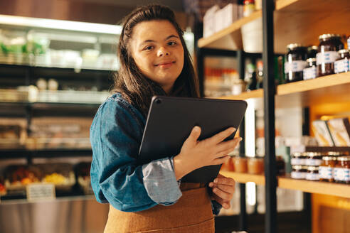 Supermarktangestellte mit Down-Syndrom, die in einem Geschäft ein digitales Tablet benutzt. Selbstbewusste Frau mit einer geistigen Behinderung, die als Verkäuferin in einem örtlichen Lebensmittelgeschäft arbeitet. - JLPSF08283