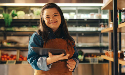 Shop employee with Down syndrome smiling at the camera while holding a digital tablet. Happy woman with an intellectual disability working as a storekeeper in a local grocery store. - JLPSF08284