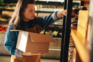 Supermarket employee with Down syndrome restocking food products onto the shop shelves. Empowered woman with an intellectual disability working as a shopkeeper in a grocery store. - JLPSF08288