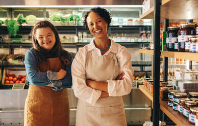 Successful grocery store workers smiling at the camera while standing together in their shop. Happy woman with Down syndrome working in a local supermarket with her female colleague. - JLPSF08292