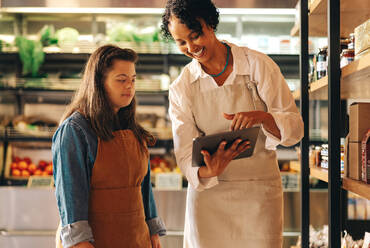 Happy grocery store manager using a digital tablet while having a discussion with her employee. Female shop owner giving a woman with Down syndrome training in managing online grocery orders. - JLPSF08293