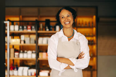 Erfolgreiche Kleinunternehmerin, die mit verschränkten Armen lächelnd am Eingang ihres Lebensmittelladens steht. Reife Unternehmerin, die einen trendigen Lebensmittelladen betreibt. - JLPSF08296
