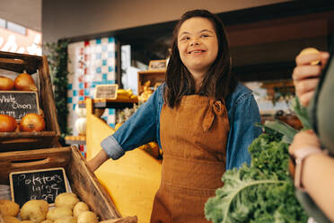 Happy woman with Down syndrome assisting a customer in the vegetable section of a grocery store. Friendly woman with an intellectual disability working as a store assistant in a supermarket. - JLPSF08304