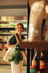 Fröhlich lächelnde Kundin, die eine Tüte mit frischem Biogemüse in einem Lebensmittelgeschäft trägt. Fröhliche junge Frau beim Einkaufen in einem trendigen Supermarkt. - JLPSF08308