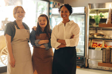 Eine Gruppe verschiedener Angestellter in einem Lebensmittelgeschäft lächelt in die Kamera. Drei glückliche Frauen arbeiten zusammen in einem erfolgreichen kleinen Unternehmen. - JLPSF08314