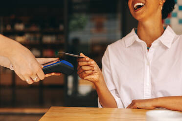 Happy female customer scanning her credit card on a card machine to pay her bill in a cafe. Cheerful woman doing a cashless and contactless transaction using NFC technology. - JLPSF08317