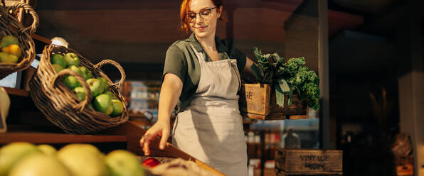 Cheerful grocery store owner restocking fresh produce in the fruit and vegetable section of her shop. Successful female entrepreneur running a small business in the food industry. - JLPSF08320