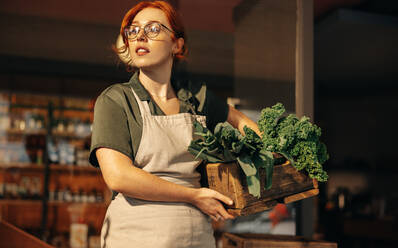 Die Besitzerin eines Lebensmittelgeschäfts schaut nachdenklich weg, während sie eine Kiste mit frischem Bio-Gemüse in ihrem Laden hält. Junge Unternehmerin, die ein kleines Unternehmen in der Lebensmittelbranche führt. - JLPSF08321