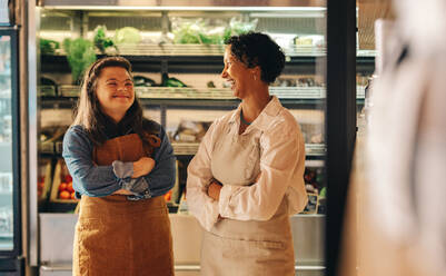 Zwei Angestellte eines Lebensmittelgeschäfts lachen fröhlich, während sie zusammen in ihrem Supermarkt stehen. Glückliche Frau mit Down-Syndrom, die zusammen mit ihrer Kollegin ein kleines Geschäft führt. - JLPSF08327
