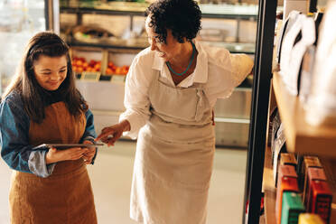 Two happy grocery store owners managing online grocery orders on a digital tablet. Empowered woman with Down syndrome running a successful supermarket with her female colleague. - JLPSF08330