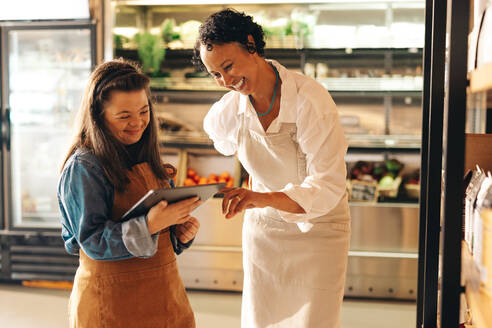 Happy grocery store workers managing online grocery orders on a digital tablet. Empowered woman with Down syndrome running a successful supermarket with her female colleague. - JLPSF08333