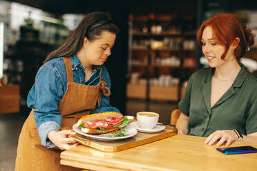 Kellnerin mit Down-Syndrom serviert einem Kunden ein Sandwich und einen Kaffee in einem trendigen Café. Berufstätige Frau mit geistiger Behinderung arbeitet in einem Schnellrestaurant. - JLPSF08334