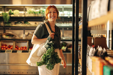 Female customer smiling cheerfully while carrying a bag with fresh organic vegetables in a grocery store. Happy young woman grocery shopping in a trendy supermarket. - JLPSF08339