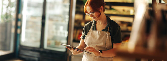 Glückliche Supermarktbesitzerin, die ein digitales Tablet benutzt, während sie in ihrem Lebensmittelgeschäft steht. Erfolgreiche Unternehmerin, die ihr kleines Unternehmen mit Hilfe der drahtlosen Technologie führt. - JLPSF08350