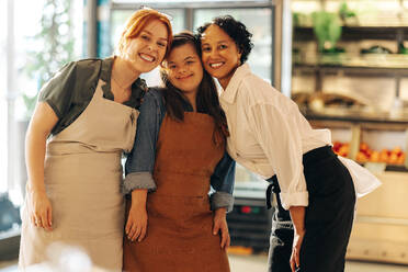 Verschiedene Einzelhandelsangestellte lächeln in die Kamera, während sie zusammen in einem Lebensmittelgeschäft stehen. Eine Gruppe von drei glücklichen Frauen, die in einem erfolgreichen Kleinunternehmen zusammenarbeiten. - JLPSF08356