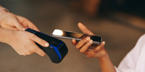 Unrecognizable customer scanning her smartphone on a credit card machine to pay her bill in a cafe. Woman doing a cashless and contactless transaction using NFC technology. - JLPSF08358