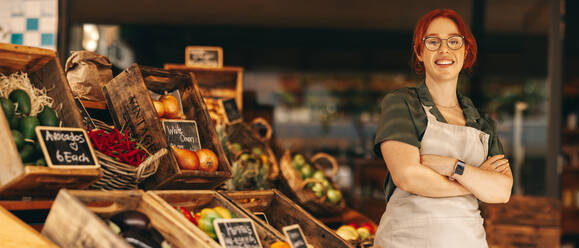 Glückliche Supermarktbesitzerin, die in die Kamera lächelt, während sie in der Frischwarenabteilung ihres Lebensmittelladens steht. Erfolgreiche junge Frau, die ein kleines Unternehmen in der Lebensmittelbranche führt. - JLPSF08362