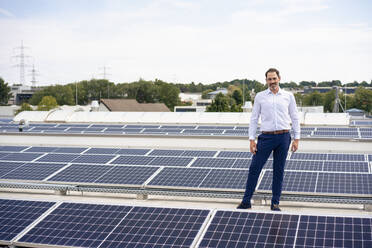Smiling businessman standing amidst solar panels on rooftop - JOSEF14409