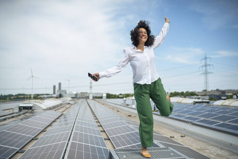 Happy businesswoman standing on one leg at rooftop with solar panels - JOSEF14438