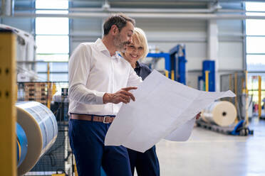 Businessman showing blueprint to smiling businesswoman in industry - JOSEF14448