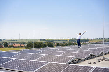 Älterer Geschäftsmann mit erhobenen Armen inmitten von Sonnenkollektoren auf dem Dach - JOSEF14462