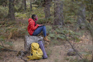 Happy woman using laptop sitting on rock in forest - JCCMF07442