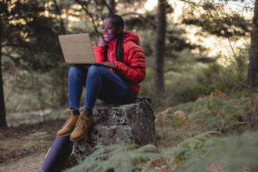 Happy young woman using laptop sitting on rock in forest - JCCMF07443