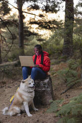 Glückliche Frau mit Laptop und Hund im Wald sitzend - JCCMF07444