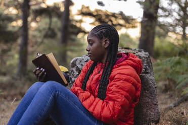 Junge Frau mit geflochtenen Haaren liest ein Buch im Wald - JCCMF07454