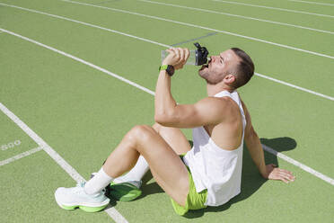 Sportler, der auf einem Sportplatz sitzend Wasser trinkt - IFRF01813