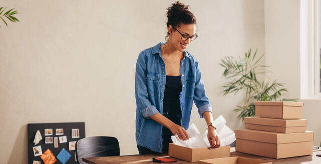 Eine Frau packt den Karton auf ihrem Schreibtisch zu Hause ein. Eine Geschäftsfrau bereitet ein Produkt für die Lieferung an einen Kunden vor. - JLPSF08398