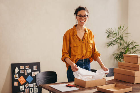Junge Frau schaut in die Kamera und lächelt im Büro des Dropshipping-Unternehmens zu Hause. Lächelnder Online-Shop-Besitzer packt Paketboxen für die Auslieferung an Kunden. - JLPSF08399